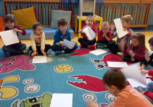 Dzieci siedzą na dywanie i machają kartkami papieru.