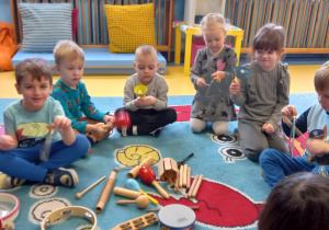 Dzieci siedzą na dywanie i swobodnie grają na instrumentach.