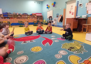Dzieci siedzą na dywanie i grają na instrumentach, dyryguje nimi dziecko - dyrygent.
