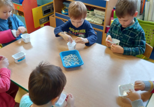 Dzieci siedzą przy stole i nakładają gumki recepturki na pojemniczki po serkach.