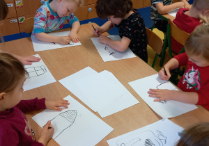 Dzieci siedzą przy stole i rysują na kartkach węglem rysunkowym.