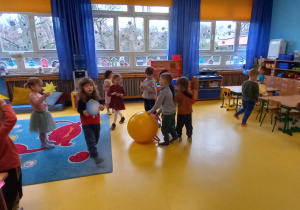 Dzieci biegają naokoło dużej żółtej piłki trzymając w rękach obrazki przedstawiające planety, inne przedszkolaki stoją trzymając sylwety gwiazd.