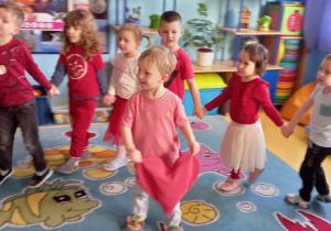 Dzieci ubrane na czerwono tańczą w kole, w środku stoi chłopiec i trzyma czerwoną poduszkę w kształcie serca.