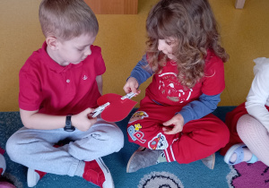 Dzieci siedzą na dywanie i podają sobie szablon serca przy pomocy spinaczy do bielizny.