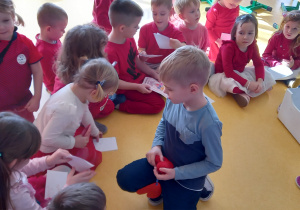 Dzieci siedzą na podłodze i podają sobie kartki walentynkowe.