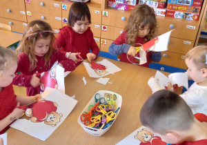 Dzieci ubrane na czerwono siedzą przy stole i wycinają sylwetkę misia z serduszkiem.