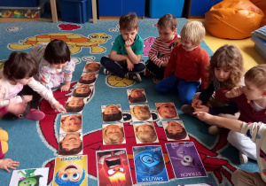 Dzieci siedzą na dywanie i dopasowują obrazki z twarzami do ilustracji przedstawiających emocje.