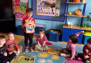 Dzieci siedzą w kole na dywanie. Chłopiec pokazuje obrazek przedstawiający radość.