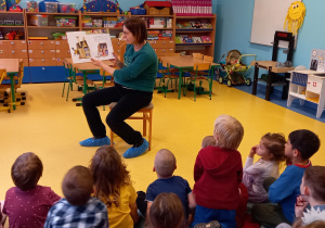 Mama chłopca z grupy siedzi na krzesełku, czyta dzieciom książkę i pokazuje ilustracje.