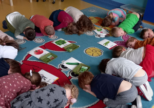Dzieci leżą skulone na dywanie w kole, w środku koła leżą tacki z nasionami warzyw i ilustracje tych warzyw.