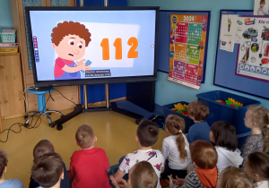 Dzieci oglądają na dużym ekranie chłopca, który dzwoni po pomoc. Na ekranie widać duże cyfry numeru 112.