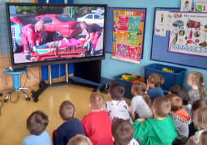 Dzieci oglądają na dużym ekranie człowieka leżącego na noszach.