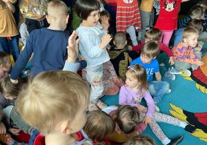 Dzieci oglądają koncert. Niektóre siedzą na dywanie, inne stoją i wykonują gesty rękami.