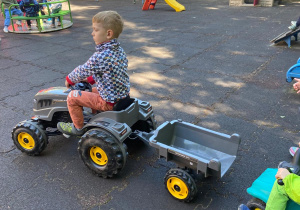 Chłopiec jedzie traktorem z przyczepką na placu zabaw.