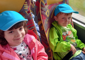 Dziewczynka i chłopiec w niebieskich czapkach z daszkiem siedzą na fotelach w autokarze.