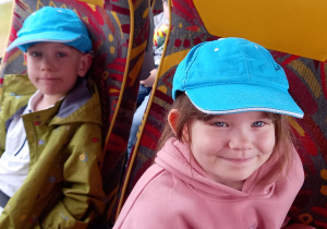 Dziewczynka i chłopiec w niebieskich czapkach z daszkiem siedzą na fotelach w autokarze.