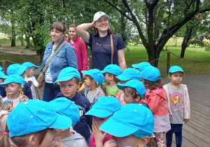 Grupa dzieci w niebieskich czapkach z daszkiem stoi w sadzie.