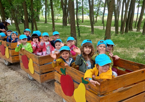 Dzieci w niebieskich czapkach z daszkiem siedzą w wagonikach kolejki.