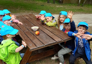 Dzieci w niebieskich czapkach z daszkiem siedzą przy stole. Na środku stołu leży jabłko.