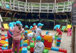 Dzieci w niebieskich czapkach z daszkiem bawią się klockami w rozmiarze XXL.