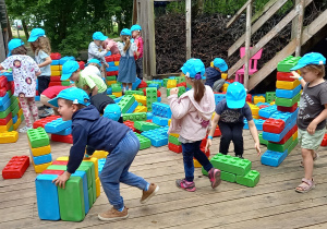Dzieci w niebieskich czapkach z daszkiem bawią się klockami w rozmiarze XXL.