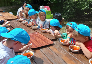 Dzieci w niebieskich czapkach z daszkiem siedzą przy stole i jedzą zupę pomidorową.
