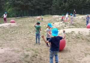Dzieci w niebieskich czapkach z daszkiem bawią się dużymi i małymi piłkami na rozległym pagórkowatym terenie.