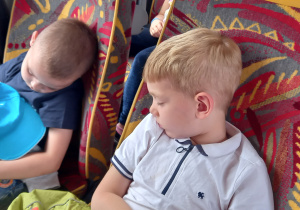 Dwóch chłopców w niebieskich czapkach z daszkiem śpią na fotelach w autokarze.