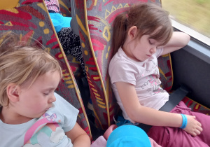 Dwie dziewczynki w niebieskich czapkach z daszkiem śpią na fotelach w autokarze.