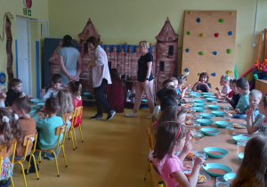 Dzieci jedzą obiad przy dwóch długich stołach, w tle widać dekorację w postaci zamku z cegieł.