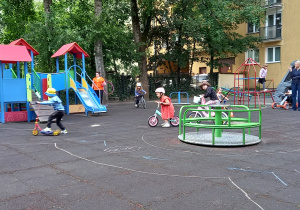 Dzieci jeżdżą na rowerkach po ścieżkach narysowanych kredą na placu zabaw.