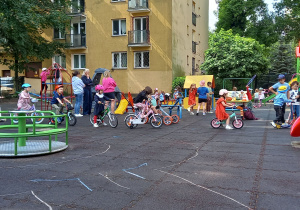 Dzieci jeżdżą na rowerkach po ścieżkach narysowanych kredą na placu zabaw.