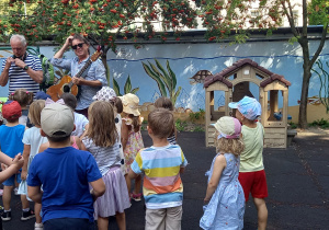 Dzieci stoją na placu zabaw i słuchają muzyka grającego na gitarze.