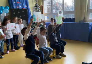 Dzień Babci i Dziadka. Czworo dzieci siedzi na krzesełkach i trzyma rysunki nad głową.