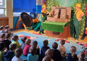 Dzieci siedzą i oglądają teatrzyk, dwoje aktorów w strojach budowlańców stoi.