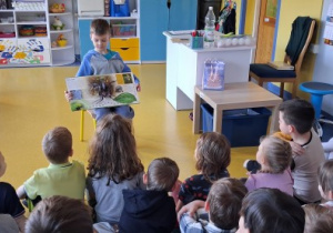 Chłopiec siedzi na krzesełku i pokazuje dzieciom swoją ulubioną książkę.
