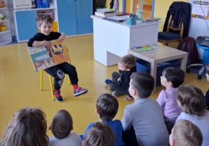 Chłopiec siedzi na krzesełku i pokazuje dzieciom swoją ulubioną książkę.