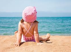 Dziewczynka w różowym kapeluszu siedzi na piasku na plaży, w tle widać niebieską morską wodę.