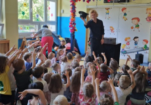 Iluzjonista pokazuje dzieciom czarodziejski kapelusz, dzieci wyciągają do niego ręce.