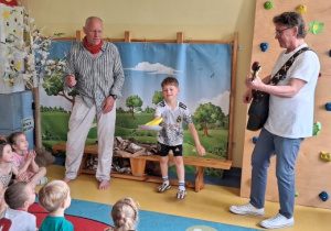 Koncert. Dzieci siedzą na dywanie, mężczyzna gra na gitarze, chłopiec podrzuca żółty talerzyk za pomocą patelni.