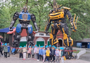Wycieczka. Grupa dzieci stoi przed dwoma wielkimi figurami robotów transformers.