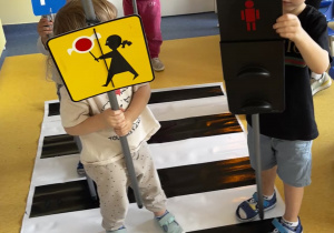 Dzieci stoją na pasach i trzymają w ręku znaki drogowe.