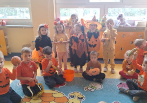 Dzieci stoją i siędzą w kręgu, są ubrane na pomarańczowo lub przebrane za dynie.