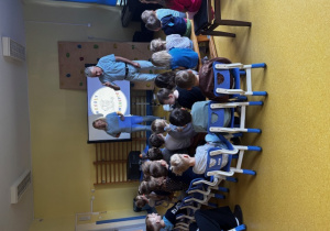 Dzieci siedzą na krzesełkach i patrzą na panią i pana w trakcie warsztatów naukowych.