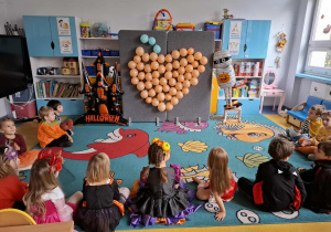 Dzieci siedzą naprzeciwko tablicy z balonami.