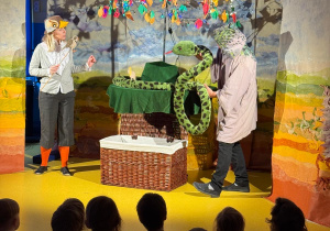Aktorzy stoją na scenie i manipulują maskotką węża.