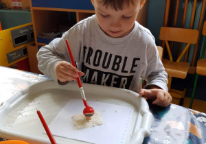 Chłopiec maluje farbą drewnianą figurkę grzyba.