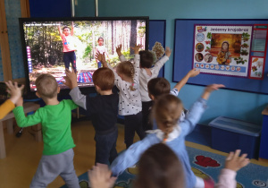 Przedszkolaki wykonują ćwiczenia razem z dziećmi na dużym ekranie.