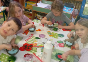 Dzieci siedzą przy stole i mieszają w miseczkach łyżeczkami niebieską substancję.